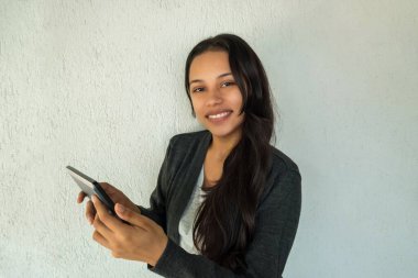 Gri ceketli genç bir kadın elinde beyaz bir arka planda tablet tutuyor. Kameraya bak..