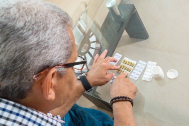 Gri saçlı yetişkin bir adam evindeki oturma odasında ilaçlarını kontrol ediyor..