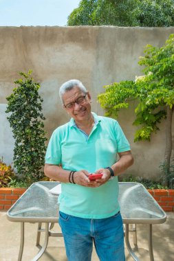 Olgun bir adam cep telefonuyla arka bahçede dikilirken kameraya bakıyor.