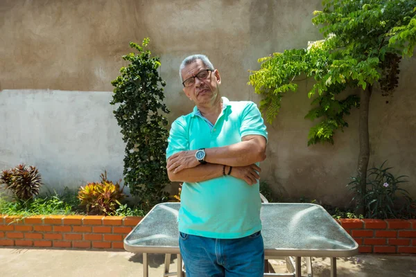 Olgun bir adam kollarını kavuşturup arka bahçede dikilirken kameraya bakıyor.