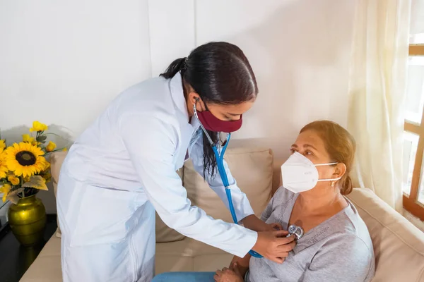 Maske takan bir doktor evde hasta olan yaşlı bir kadının kalp atışlarını kontrol etmek için steteskop kullanıyor.