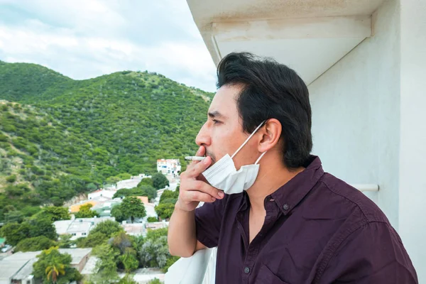 30-year-old man with mask on the balcony of his house smoking a cigarette