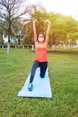 Maskeli kadın yoga pozu verirken parkta bir yatakta bacağını esnetiyor. Yeni normallik kavramı