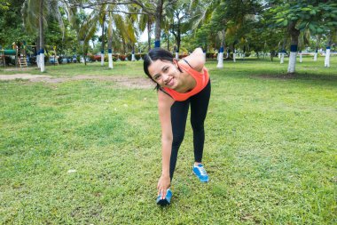 Park çimenliğinde kameranın önünde gülümseyen ve gerinen bir kadın.