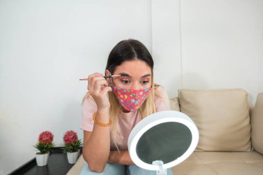 Coronavirus koruması takan kadın yüz maskesi ve makyaj. Maskeli bir kadın aynada kendine bakıyor ve makyaj yapıyor. Tıbbi Maske, Yakın çekim, COVID-19 Önleme