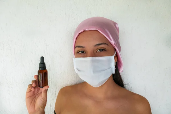 Latin woman with medical mask and pink scarf holding cosmetic oil for facial skin care