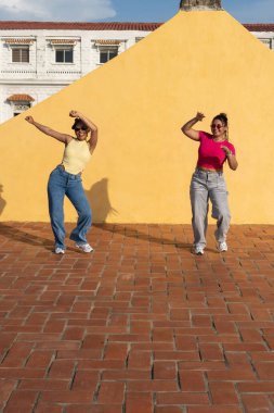 İki genç kadın, canlı sarı bir duvara karşı çatıda mutlu bir şekilde dans ediyor.