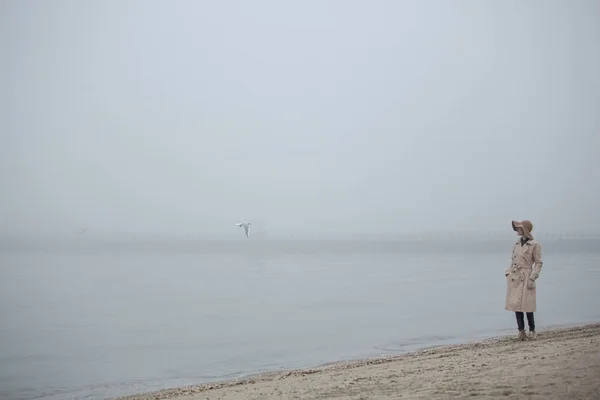 Foggy morning at sea. Woman in hat and coat walking on autumn beach