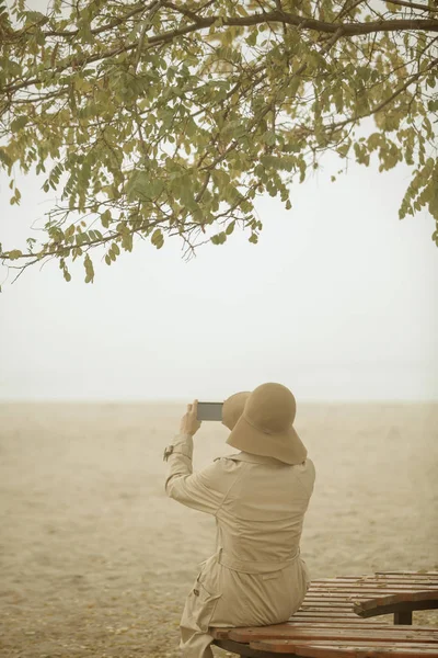 若い女性の女性の Wwearing の帽子とコートを秋風景孤独な秋ビーチのスマート フォンを使用しての背面図 — ストック写真