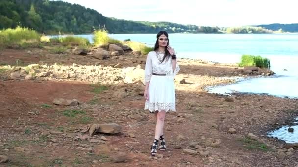 एक पांढरा ड्रेस आणि जातीय दागिने सुंदर मुलगी नदीच्या काठावर कॅमेरावर चालत आहे — स्टॉक व्हिडिओ