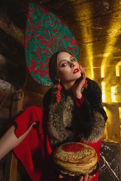 Uma menina com panquecas e caviar, em um kokoshnik vermelho e casaco preto, senta-se em um celeiro . — Fotografia de Stock