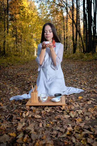En flicka i blå kimono sitter i höstskogen och förbereder te — Stockfoto
