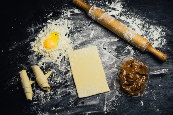 Ingredients and utensils for the preparation of bakery products - flour, dough, egg, rolling pin, whisk, strainer, bread - on black table with free copy space