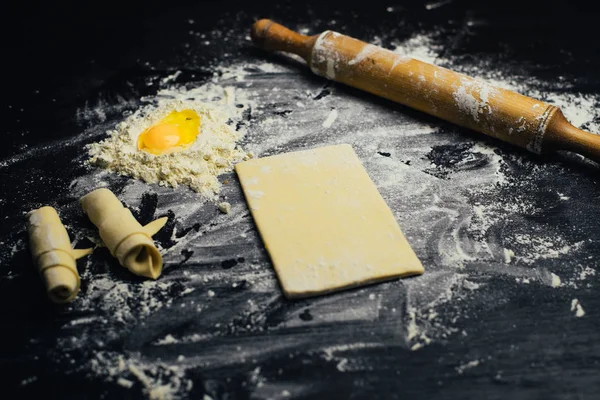 Ingredients and utensils for the preparation of bakery products - flour, dough, egg, rolling pin, whisk, strainer, bread - on black table with free copy space