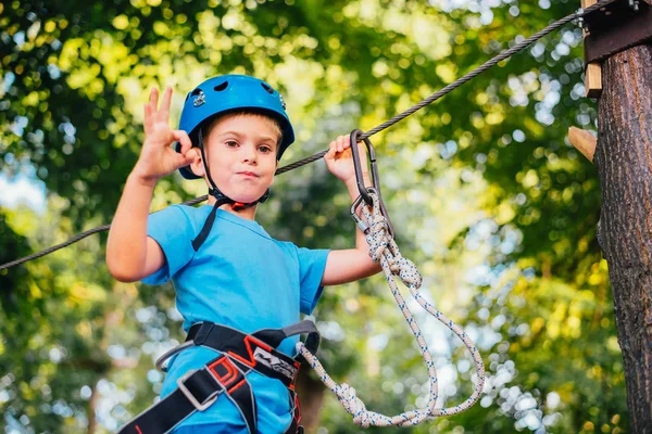 Little cute boy enjoying activity in a climbing adventure park on a summer sunny day.  Climbing extreme sport with helmet and insurance