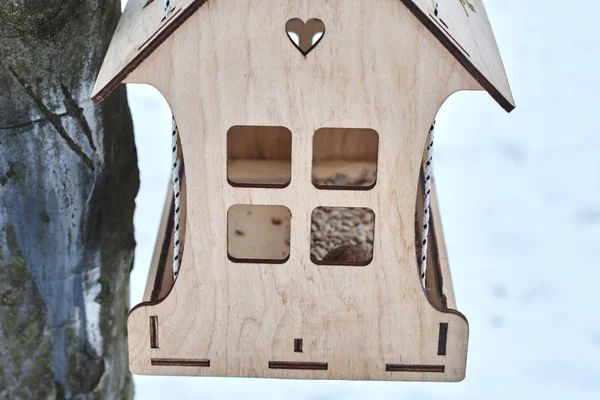 Stylish bird feeder made with plywood sheets filled with seeds hanging on tree in winter forest