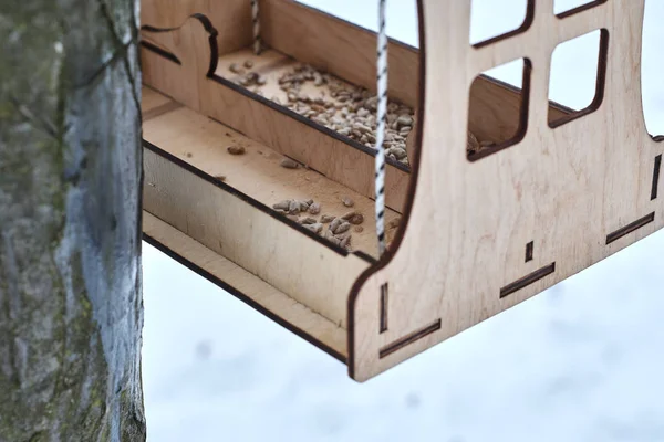 Stylish bird feeder made with plywood sheets filled with seeds hanging on tree in winter forest