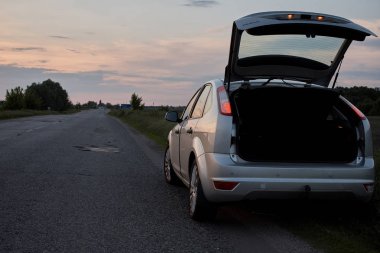 Bulutlu gün batımında kırsal yolda park edilmiş gümüş renkli parlak araba.  