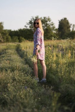 Çiçeklerle tarlada poz veren, elbiseli ve güneş gözlüklü güzel bir kadın.