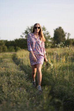 Çiçeklerle tarlada poz veren, elbiseli ve güneş gözlüklü güzel bir kadın.