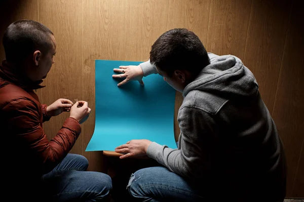back view of men crouching on the floor with blank blue paper near wooden wall