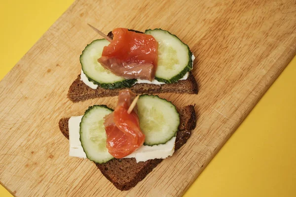 Sandwiches with salmon and cucumbers served on wooden board on yellow background