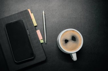 Masasında defter, akıllı telefon ve kalemle birlikte bir fincan kahve. Çalışma alanı konsepti, üretkenlik ve planlama geçmişi