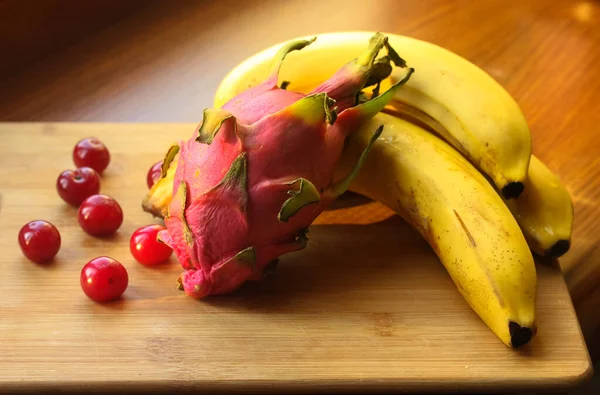 Banane, ciliegie e pitaie, frutta del drago: frutti maturi e succosi giacciono su una tavola di legno su un tavolo.. — Foto Stock