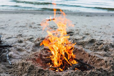 Kumlu bir plajda kamp ateşi alevleri ile sıcaklığı ve rahatlığı davet eder. Okyanus kıyısında bir şömine ışıl ışıl yanar. Alevler arka plandaki sakin suya ulaşır..