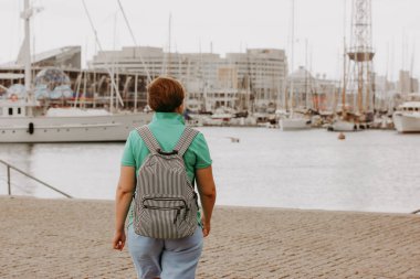 Sırt çantalı olgun bir kadın Barcelona 'da liman manzarasına bakıyor. Çizgili sırt çantalı bir kadın, Barcelona, İspanya' da tekne ve yatlarla dolu bir yat limanına bakan bir rıhtımda yürüyor..