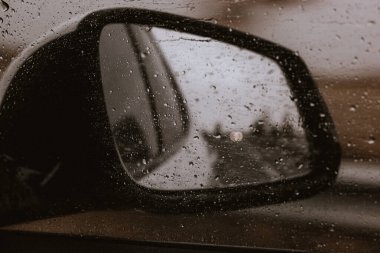 Bir arabanın yan aynasında yağmur damlaları yağmurlu bir günde karamsar, atmosferik bir manzara yaratır. Yağmur damlalarıyla kaplı bir arabanın yan aynasının yakın çekimi, bulanık bir açık hava sahnesini yansıtıyor. Yağmurlu bir araba gezisi. 