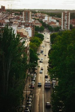Madrid, İspanya 1 Mayıs 2025 Arabalı ve yemyeşil ağaçlı bir şehir caddesinin havadan görünüşü. Bir tepeden çekim, yol boyunca hareket eden arabalar ve kamyonlarla ağaçlarla ve binalarla dolu bir şehir caddesini yakalıyor.