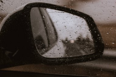 Bir arabanın yan aynasında yağmur damlaları yağmurlu bir günde karamsar, atmosferik bir manzara yaratır. Yağmur damlalarıyla kaplı bir arabanın yan aynasının yakın çekimi, bulanık bir açık hava sahnesini yansıtıyor. Yağmurlu bir araba gezisi. 