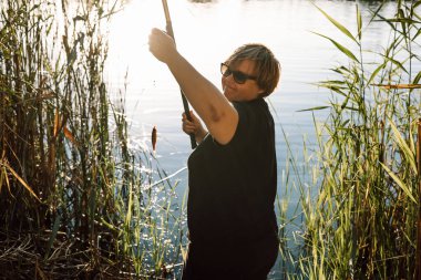 Güneşli bir günde oltaya takılmış küçük bir balıkla balık tutan mutlu olgun bir kadın. Güneş gözlüğü takan biri oltaya takılmış bir balıkla balık tutuyor. Arka planda sazlıklar ve göl görünüyor..