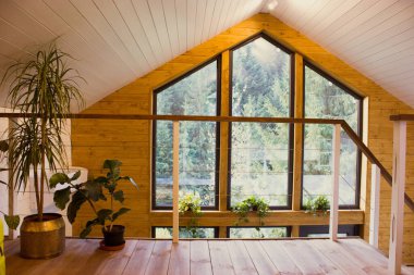 Geniş pencereli, yemyeşil bir ormana bakan rahat, ahşap bir kulübe. Orman manzarası, bitkiler ve merdiveni olan büyük bir pencereye sahip ahşap bir kulübe manzarası..