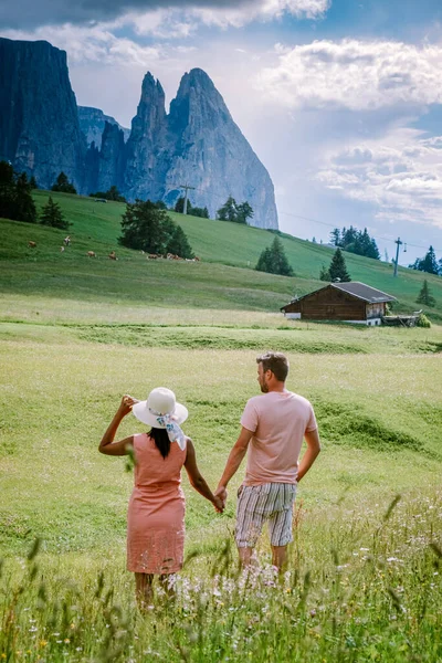 Пара мужчин и женщин на отдыхе в Доломитовых Альпах Италии, Альпе ди Сюзи - Сейзер Альм с Сассолунго - Лангкофель горной группы на заднем плане на закате. Желтые весенние цветы и деревянные шале в — стоковое фото