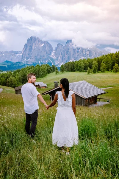 Paar im Urlaub in den Dolomiten Italien, Seiser Alm mit Langkofel-Langkofelgruppe im Hintergrund bei Sonnenuntergang. Gelbe Frühlingsblumen und Holzchalets in — Stockfoto