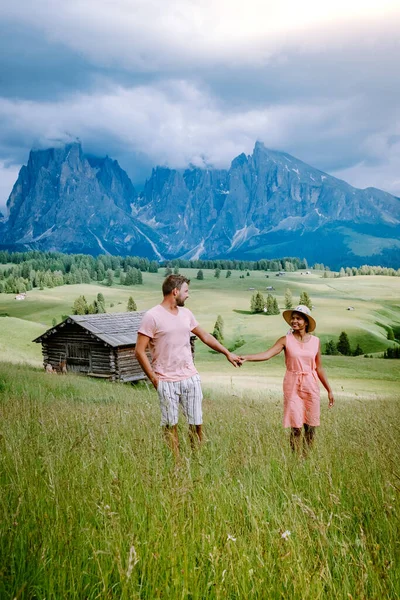 Paar im Urlaub in den Dolomiten Italien, Seiser Alm mit Langkofel-Langkofelgruppe im Hintergrund bei Sonnenuntergang. Gelbe Frühlingsblumen und Holzchalets in — Stockfoto