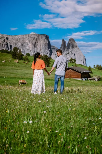 Paar mannen en vrouwen op vakantie in de Dolomieten Italië, Alpe di Siusi - Seiser Alm met Sassolungo - Langkofel berggroep op de achtergrond bij zonsondergang. Gele lentebloemen en houten chalets in — Stockfoto