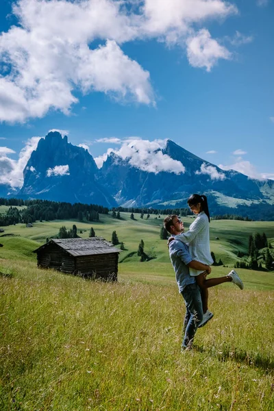 Paar mannen en vrouwen op vakantie in de Dolomieten Italië, Alpe di Siusi - Seiser Alm met Sassolungo - Langkofel berggroep op de achtergrond bij zonsondergang. Gele lentebloemen en houten chalets in — Stockfoto