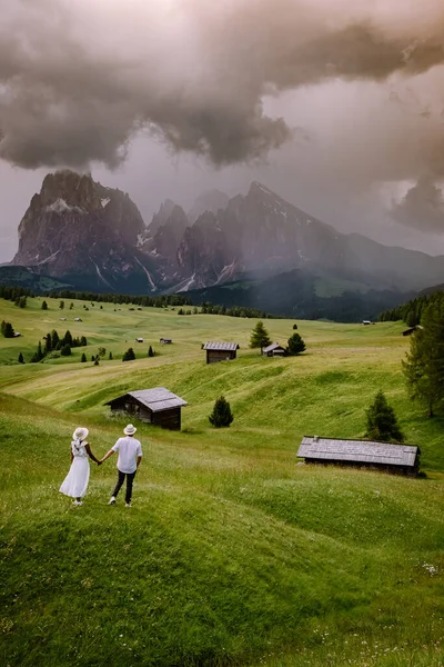 Ζευγάρι ανδρών και γυναικών σε διακοπές στο Dolomites Ιταλία, Alpe di Siusi - Seiser Alm με Sassolungo - Langkofel ορεινή ομάδα στο παρασκήνιο κατά το ηλιοβασίλεμα. Κίτρινα ανοιξιάτικα λουλούδια και ξύλινα σαλέ — Φωτογραφία Αρχείου