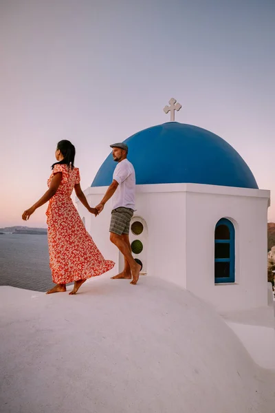 Santorini Grecia, pareja joven en vacaciones de lujo en la isla de Santorini viendo el amanecer por la iglesia de cúpula azul y el pueblo encalado de Oia Santorini Grecia durante el amanecer, hombres y mujeres en — Foto de Stock