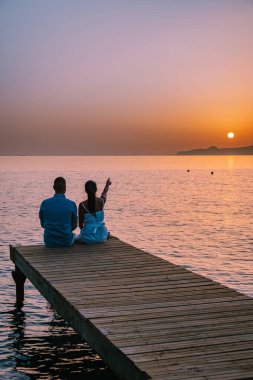 Girit Yunanistan 'da, birbirine aşık genç romantik çift gün doğumunda altın gökyüzü eşliğinde kumsaldaki ahşap iskelede oturup sarılıyor. Tatil ve seyahat konsepti. Deniz kenarında flört eden romantik genç çift
