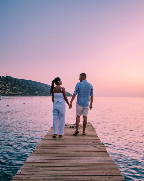 Girit Yunanistan 'da, birbirine aşık genç romantik çift gün doğumunda altın gökyüzü eşliğinde kumsaldaki ahşap iskelede oturup sarılıyor. Tatil ve seyahat konsepti. Deniz kenarında flört eden romantik genç çift — Stok fotoğraf