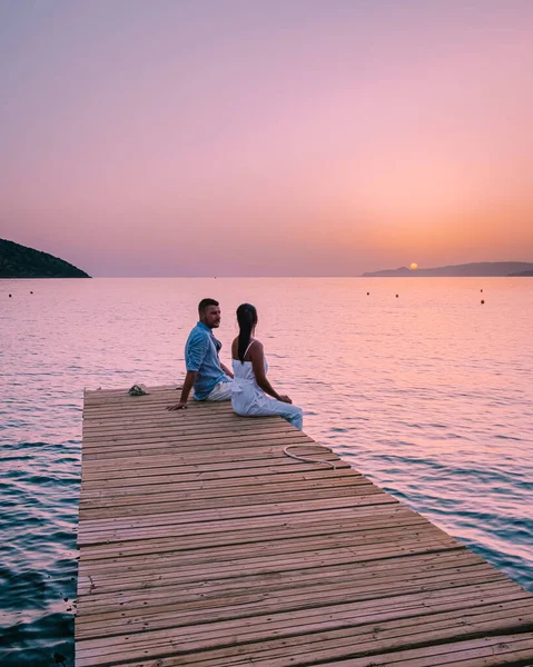 Crete Greece, young romantic couple in love is sitting and hugging on wooden pier at the beach in sunrise time with golden sky. Vacation and travel concept. Romantic young couple dating at seaside — Stock Photo, Image