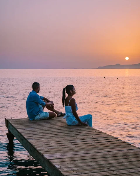 クレタ島ギリシャ、愛の若いロマンチックなカップルは黄金の空と日の出の時間にビーチで木製の桟橋に座っていると抱擁しています。休暇と旅行の概念。ロマンチックな若いカップル日付海辺で — ストック写真