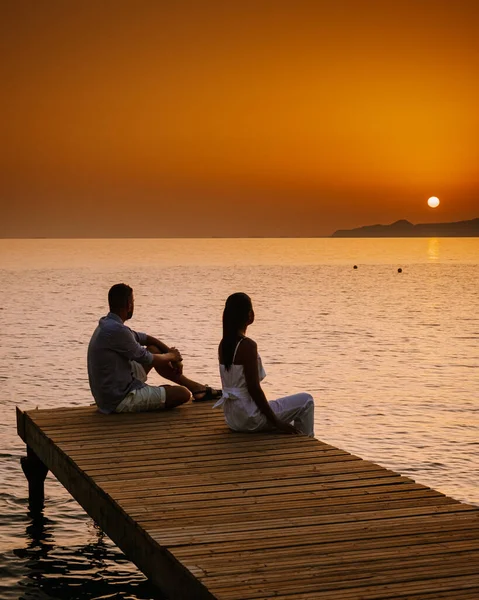 Crete Greece, young romantic couple in love is sitting and hugging on wooden pier at the beach in sunrise time with golden sky. Vacation and travel concept. Romantic young couple dating at seaside — Stock Photo, Image