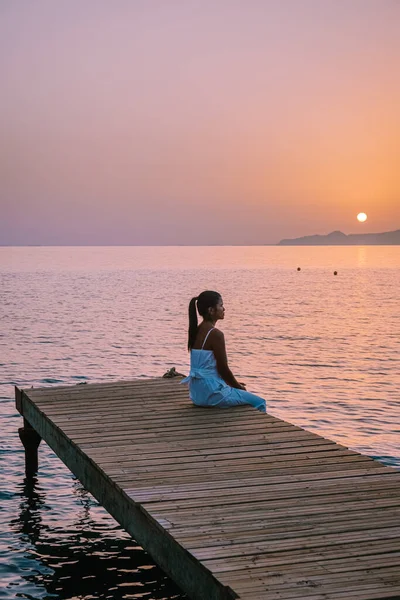 Creta Grecia, joven pareja romántica enamorada se sienta y se abraza en el muelle de madera en la playa en el amanecer con el cielo dorado. Concepto de vacaciones y viajes. Romántica pareja joven citas en la playa — Foto de Stock