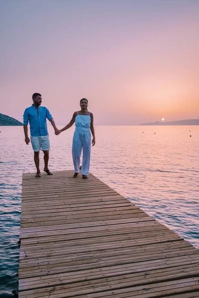 クレタ島ギリシャ、愛の若いロマンチックなカップルは黄金の空と日の出の時間にビーチで木製の桟橋に座っていると抱擁しています。休暇と旅行の概念。ロマンチックな若いカップル日付海辺で — ストック写真