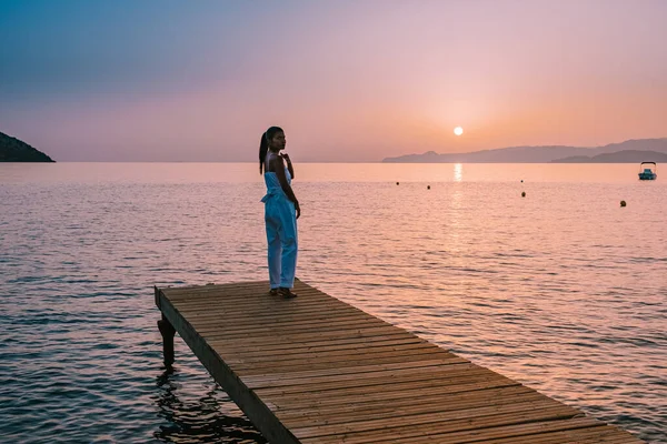 Creta Grecia, joven pareja romántica enamorada se sienta y se abraza en el muelle de madera en la playa en el amanecer con el cielo dorado. Concepto de vacaciones y viajes. Romántica pareja joven citas en la playa — Foto de Stock